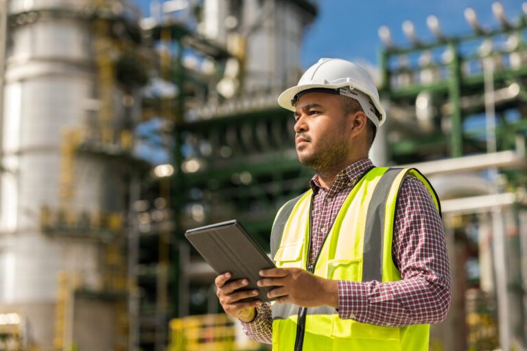 Asian engineer handsome man use tablet with white safety helmet standing front of oil refinery. Industry zone gas petrochemical. Factory oil storage tank and pipeline. Workers in a refinery