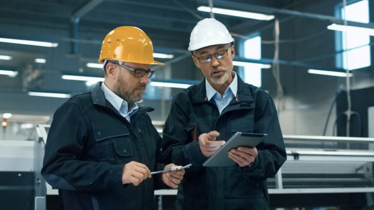 Two engineers in hardhats discuss information on a tablet computer while standing in a factory.