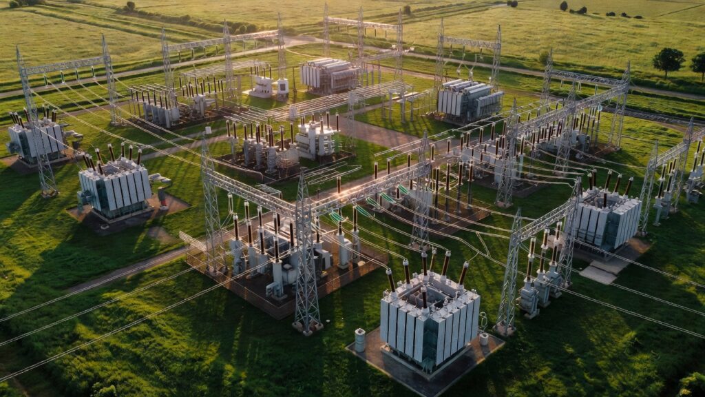 Aerial view of an electrical substation with large transformers and power lines, set in a green grassy field with roads and distant trees, photographed at sunset.